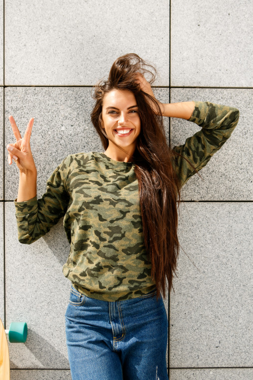 Happy beautiful woman with long sexy healthy hair in khaki top and ripped jeans with wooden longboard skateboard look to camera, show victory sign and smile. Urban scene, city life. Hipster cute lady.